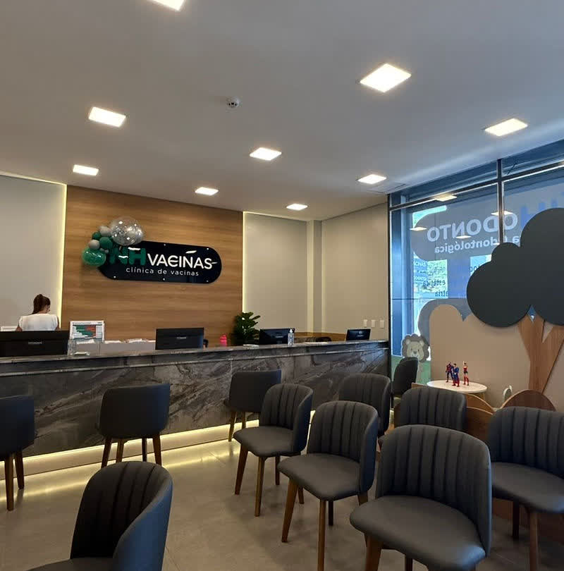 Recepção da NH Vacinas com balcão de madeira, logo NH VACINAS, cadeiras cinza e área infantil